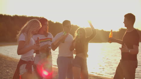 Group-of-students-celebrate-graduation-with-beer-on-the-sand-beach.-They-are-dancing-on-the-open-air-party-at-sunset.